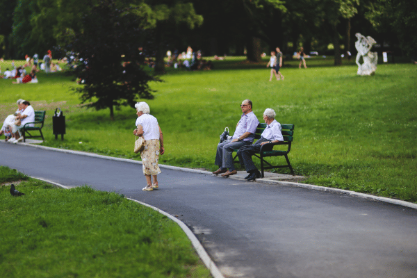 Elderly people are sitting and walking in a peaceful green park with benches and trees around.